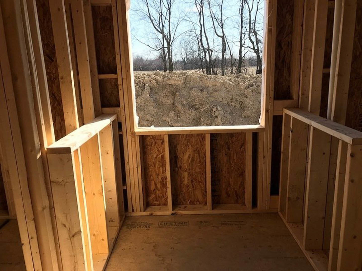Bright room featuring a large window framed in wood, offering a view of leafy trees outside; light hardwood floors and smooth plank walls enhance the natural, airy atmosphere.
