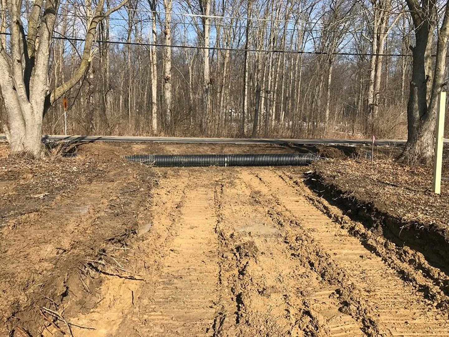 Dirt road with tire tracks, black pipe lying on soil, surrounded by trees and power lines in a wooded area