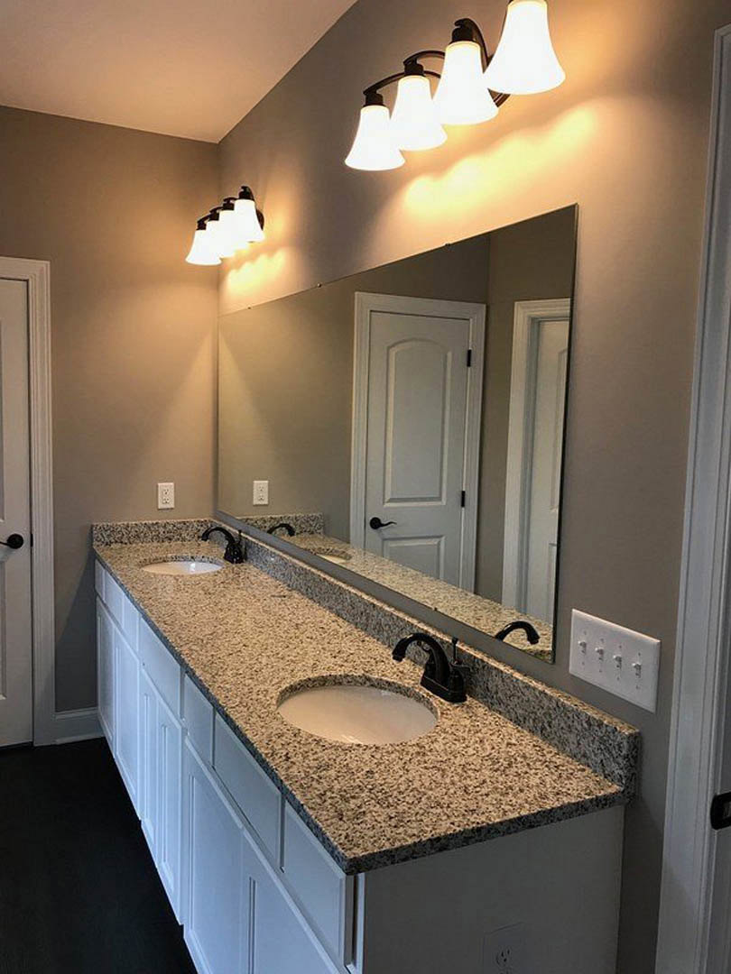 Bathroom with double vanity featuring undermount sinks, quartz countertop, expansive framed mirror, row of modern wall-mounted light fixtures, white cabinetry, and white door with
