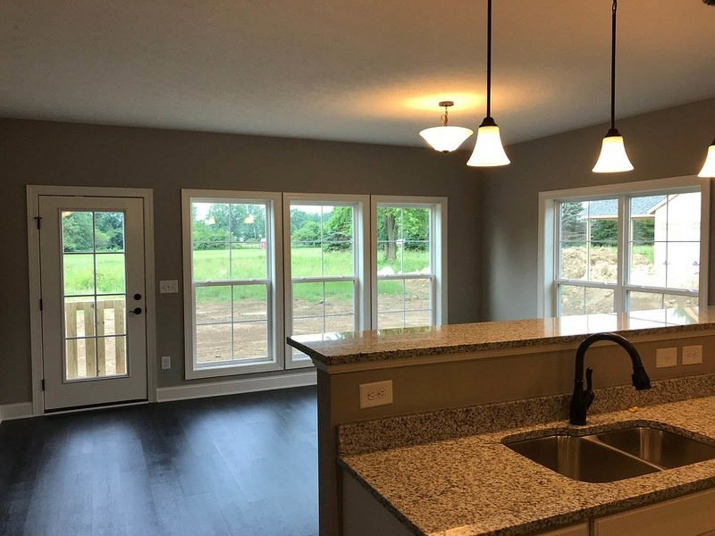 Open kitchen and living room featuring large windows overlooking grassy yard, tile backsplash, stone countertops, stainless steel sink and faucet, glass-paneled door, dark wood