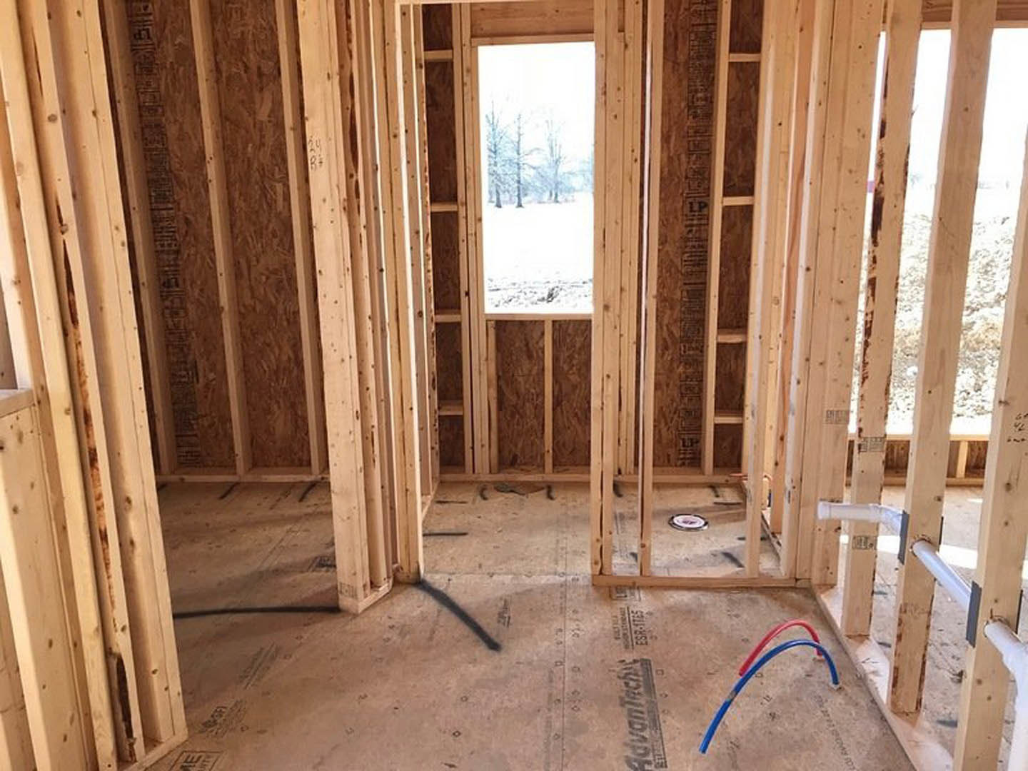 Wood-framed room under construction with exposed beams, blue and red plumbing pipes, white insulation pipes, large window overlooking snowy trees