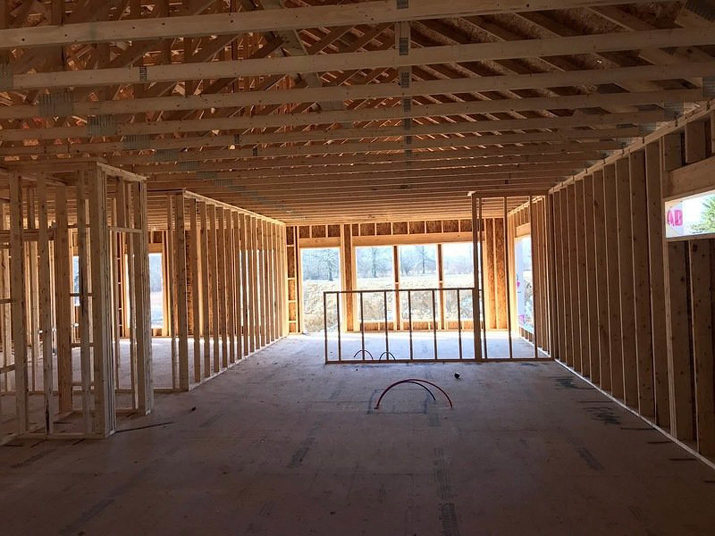 Framed room with exposed wood beams, large window, concrete floor with visible metal pipes, and unfinished ceiling