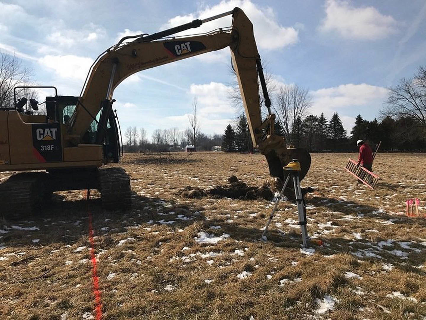 Yellow bulldozer parked on grassy field with patches of snow, surrounded by trees under a cloudy sky