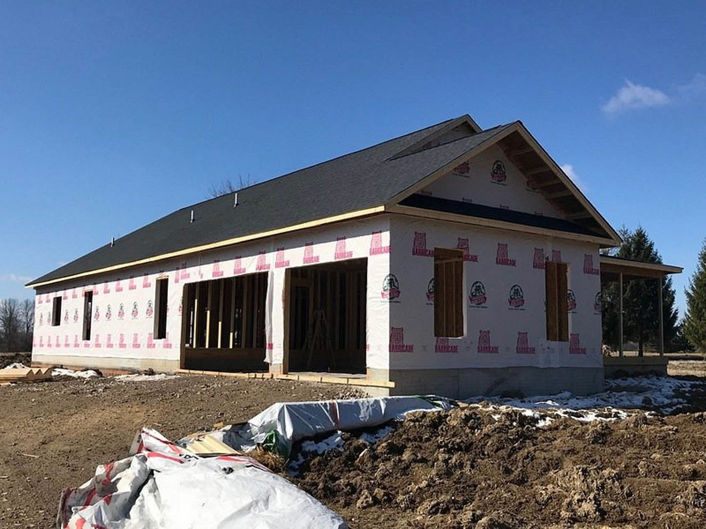 Wood-framed house under construction with exposed sheathing, pile of dirt in foreground, scattered white plastic bags, red caution tape, and clear blue sky overhead