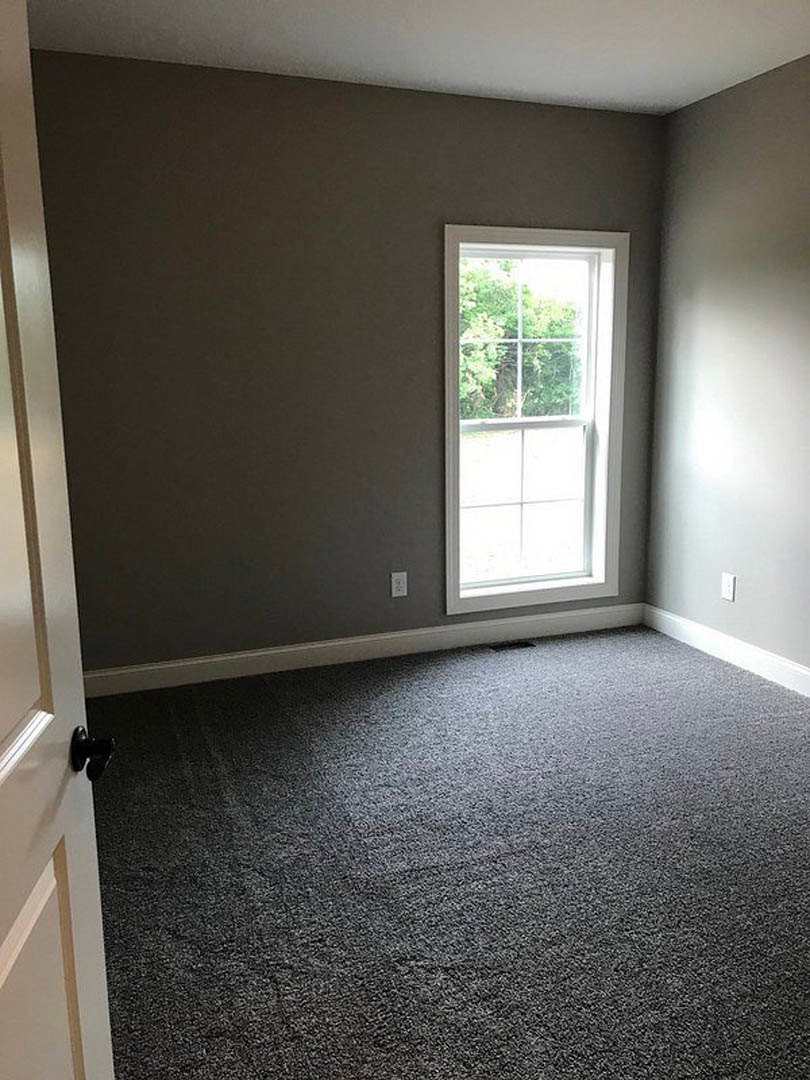 Bright room with white walls, large window framed by white trim, view of green trees outside, soft beige carpet flooring, black door knob on white door, smooth plaster ceiling