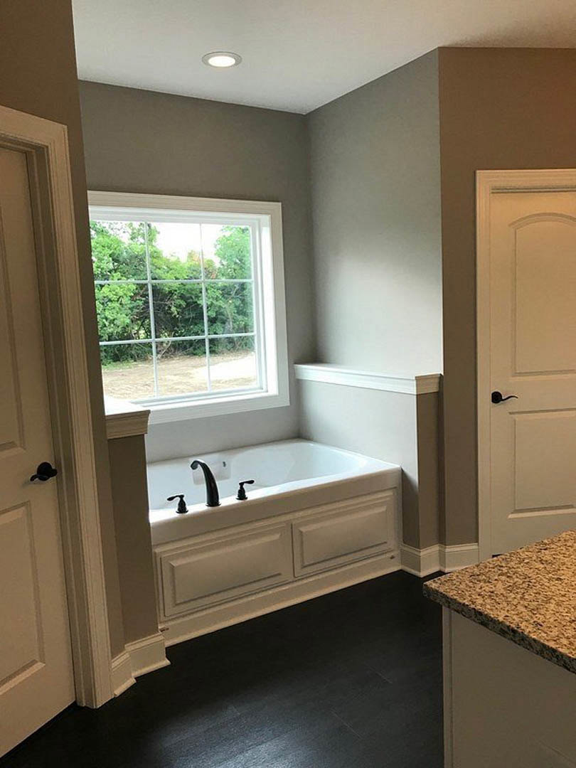 Freestanding white bathtub with black faucet and handle, multi-pane window, light tile floor, white cabinetry, and stone countertop in a modern bathroom