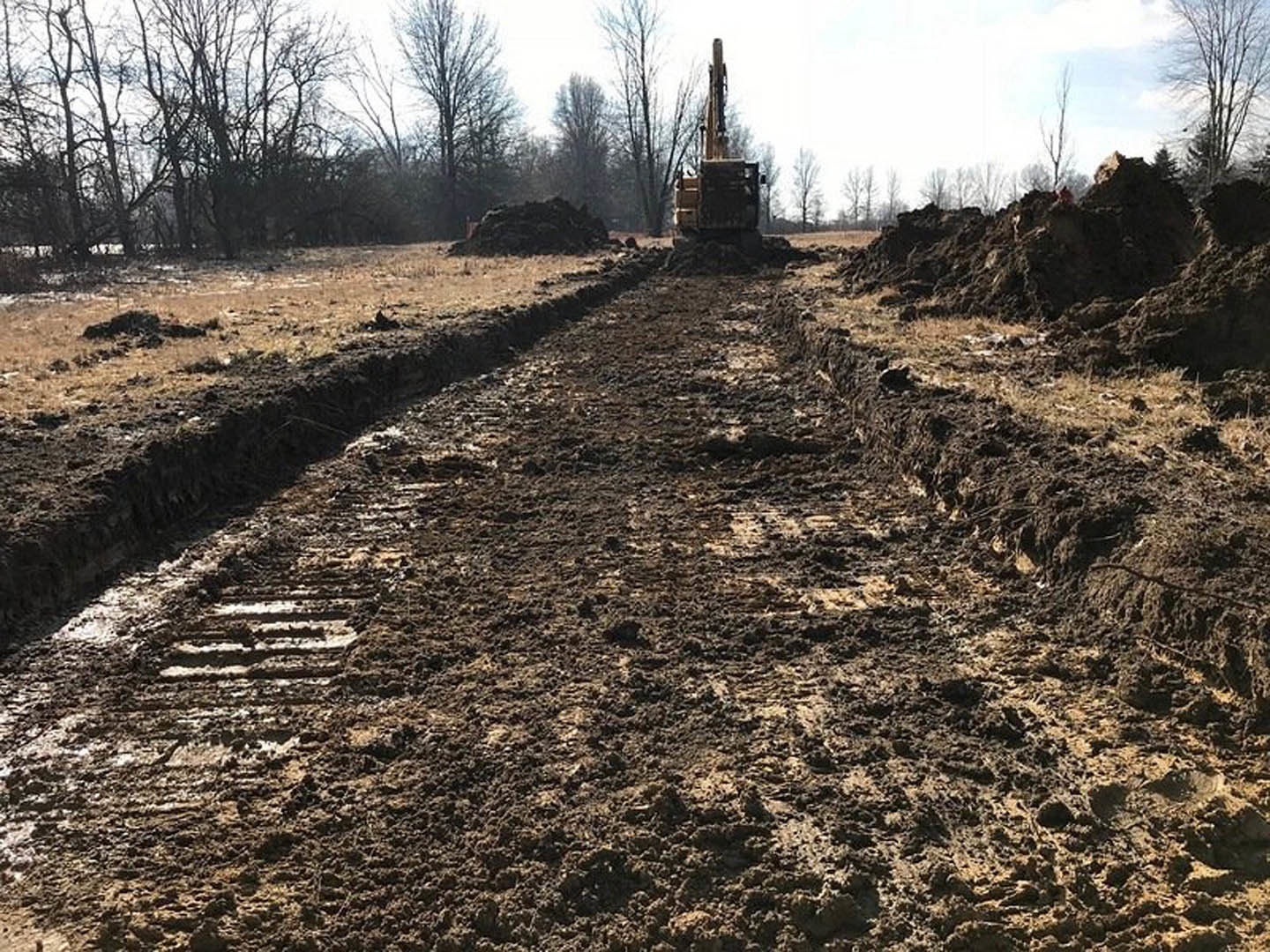 Bulldozer parked on a muddy dirt road surrounded by trees and grass, tire tracks visible in soil