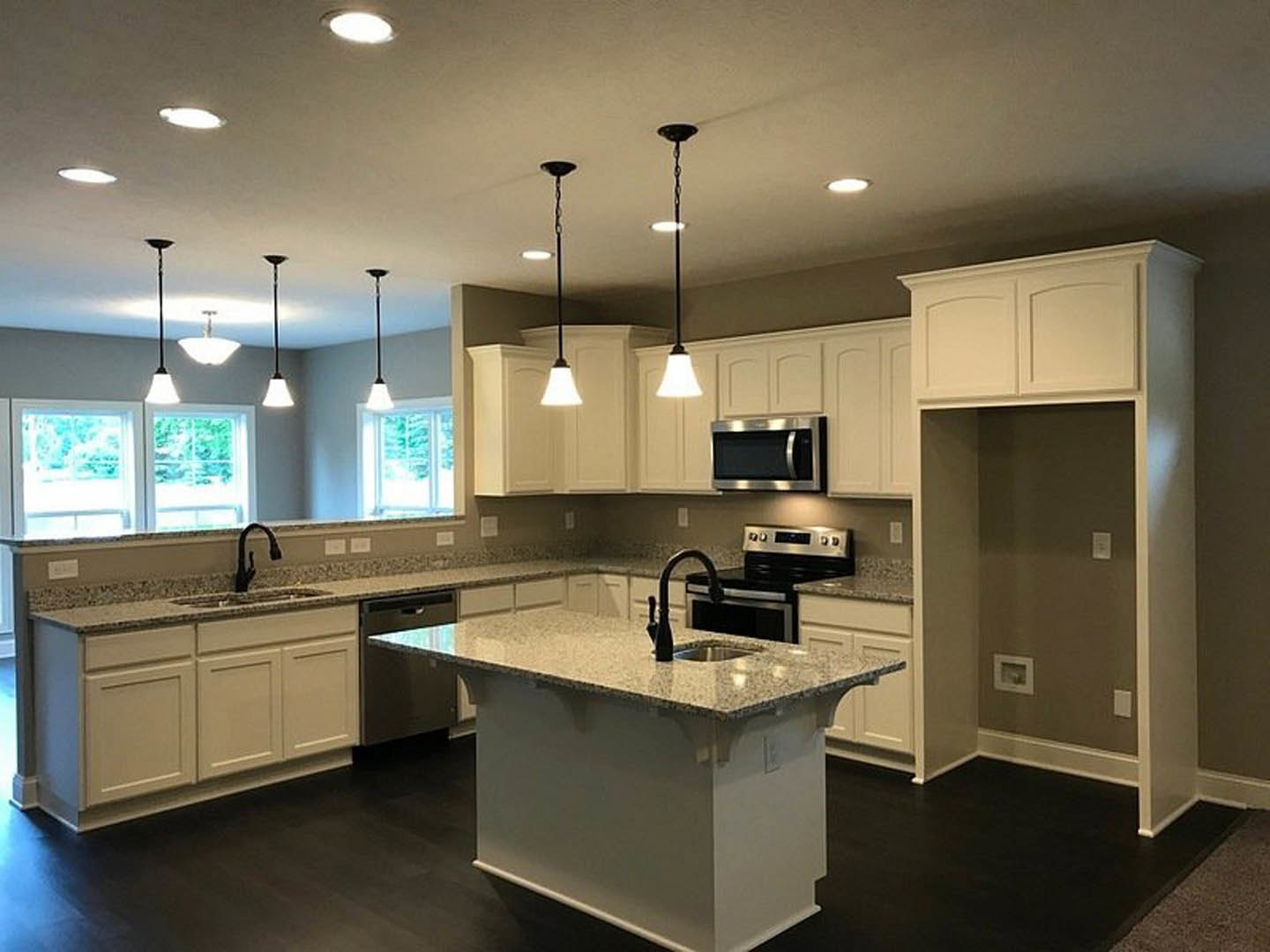 White kitchen with granite countertops, stainless steel microwave and stove, central island with built-in sink, tile backsplash, recessed ceiling light, and white cabinetry.