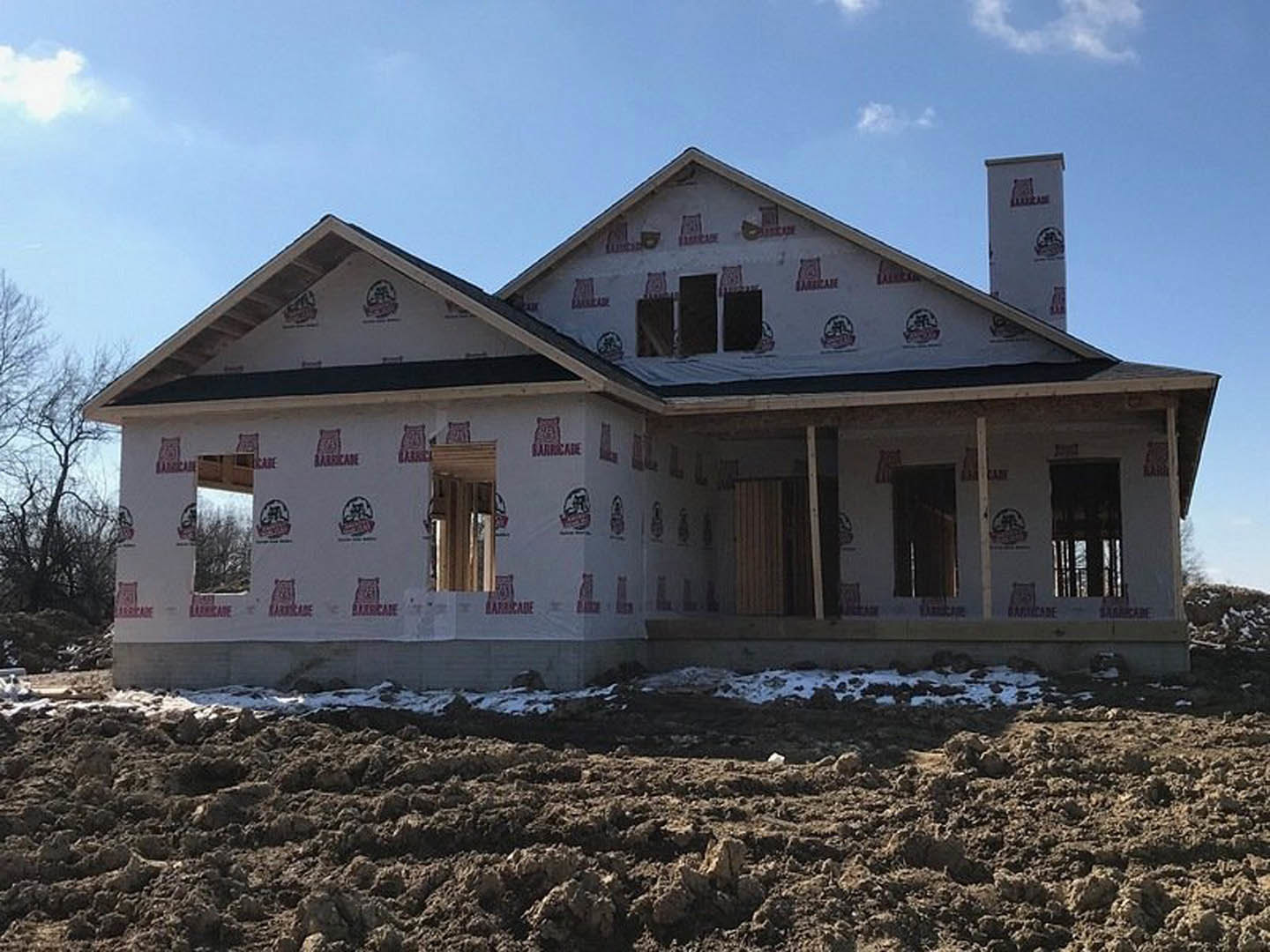 Framed house under construction with white sheeting, exposed dirt lot, leafless tree, and blue sky with scattered clouds