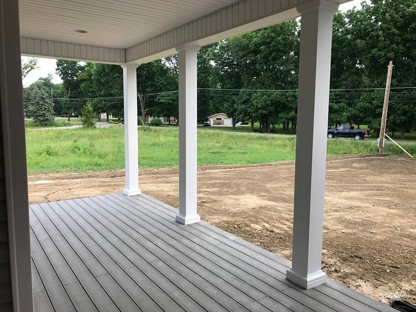 Wooden porch with white pillars overlooking a dirt field, grass and trees in the background, shaded outdoor area with windows visible on the building.