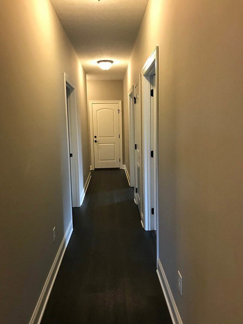 Hallway with white paneled doors featuring black knobs, dark wood flooring with white baseboards, white walls, and a wall-mounted light fixture