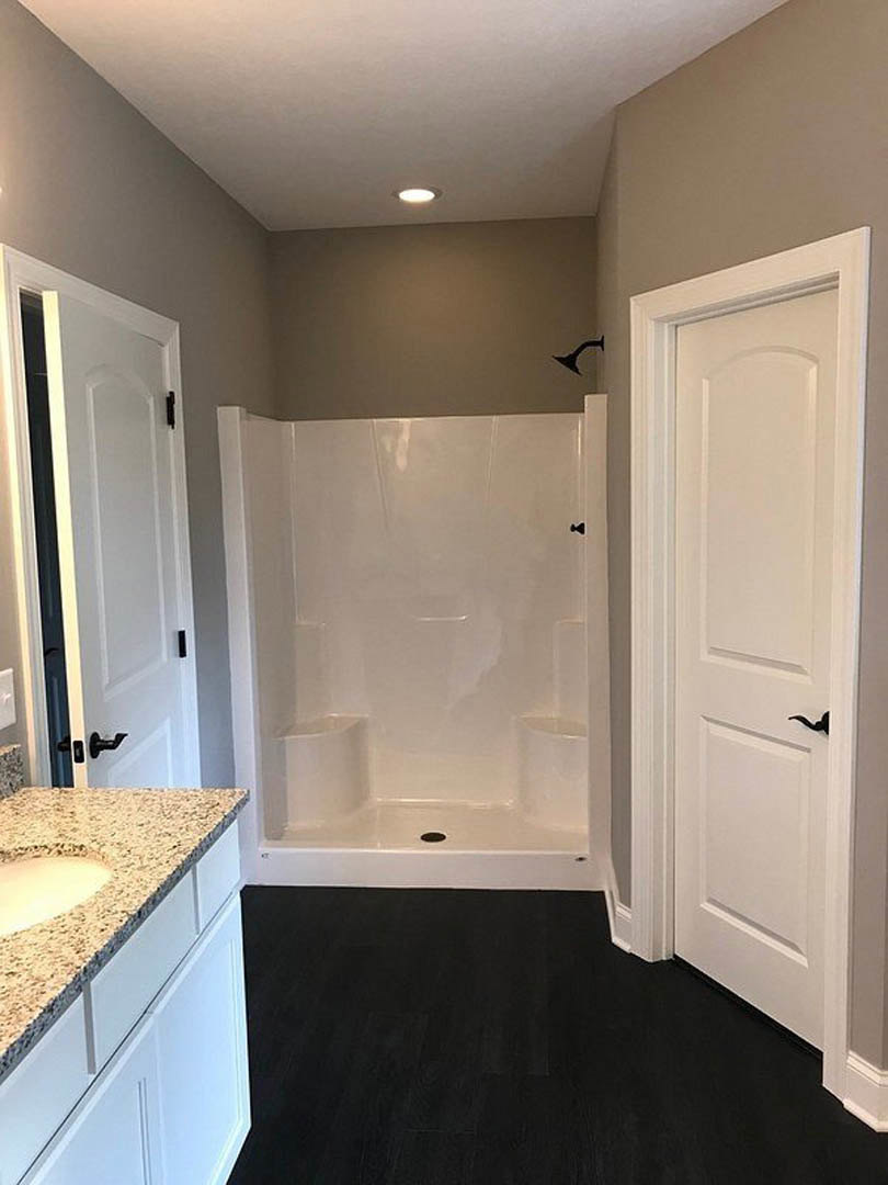 Bathroom with glass-enclosed white tile shower, marble countertop vanity, black faucet, white cabinetry, black wood floor, and white door with black handle