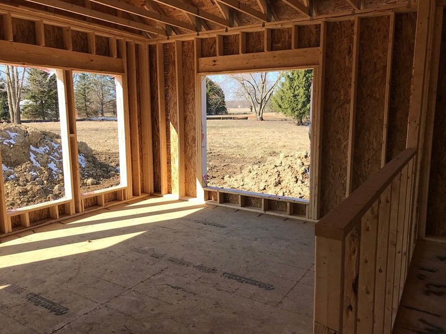Sunlit room with wood plank flooring, wooden railing, large window overlooking dirt field and trees, window frame casting shadow on ground.