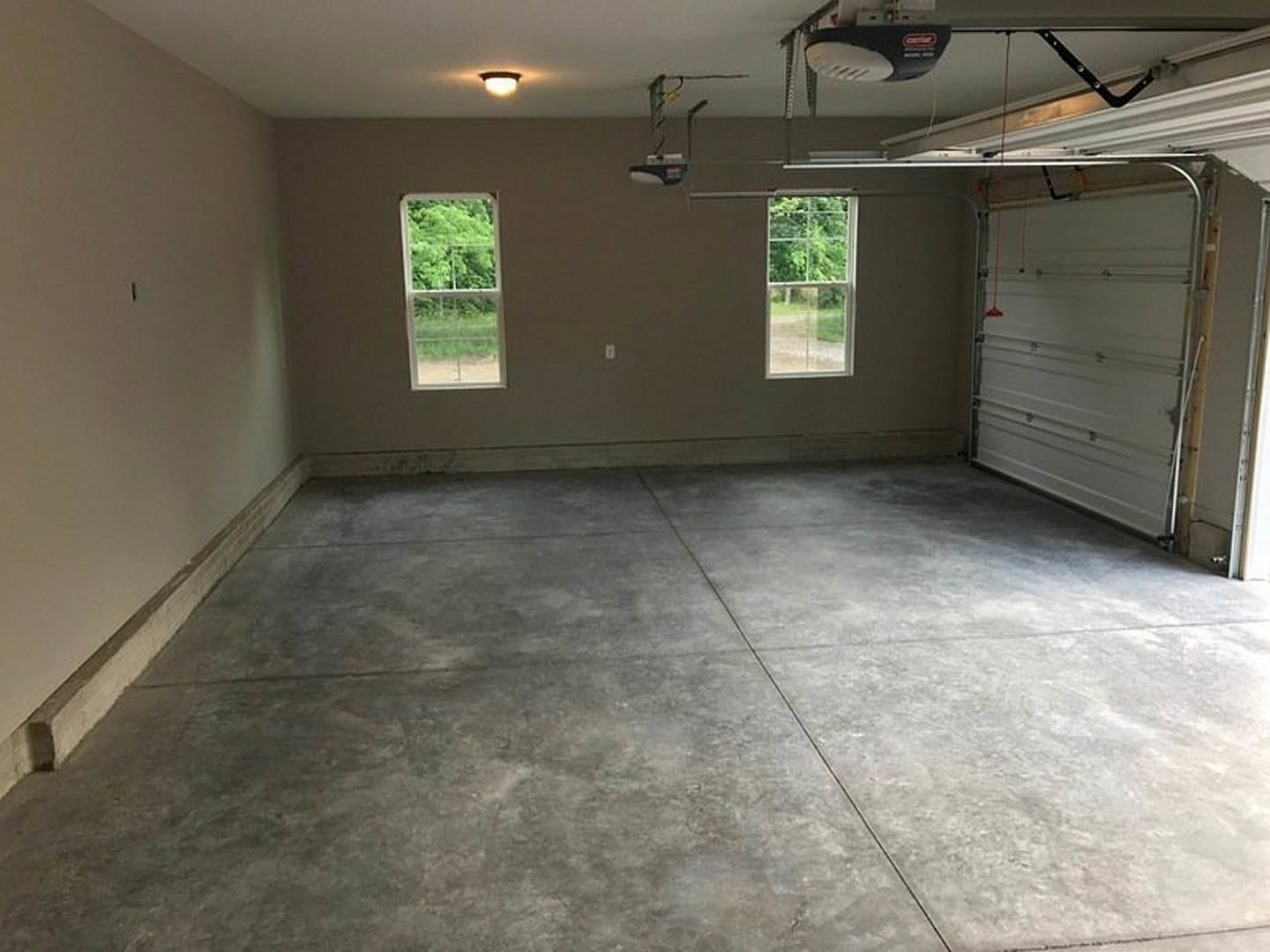 Spacious garage with grey tile flooring featuring a central crack, white plaster walls, large garage door, and windows overlooking trees
