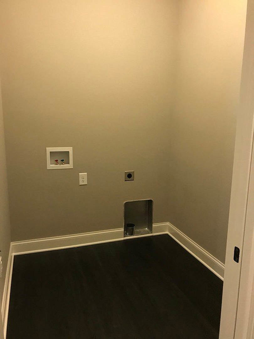 Small bathroom with black flooring, white plaster walls, white shelving unit with metal frame, silver bin recessed in floor corner, and dark wood floor accents