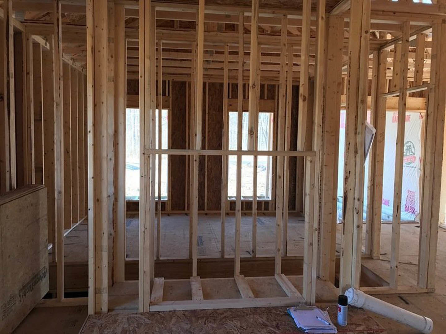 Exposed wood framing with beams and studs, white plumbing pipe on subfloor, insulation visible between lumber, construction materials and signage attached to unfinished wall