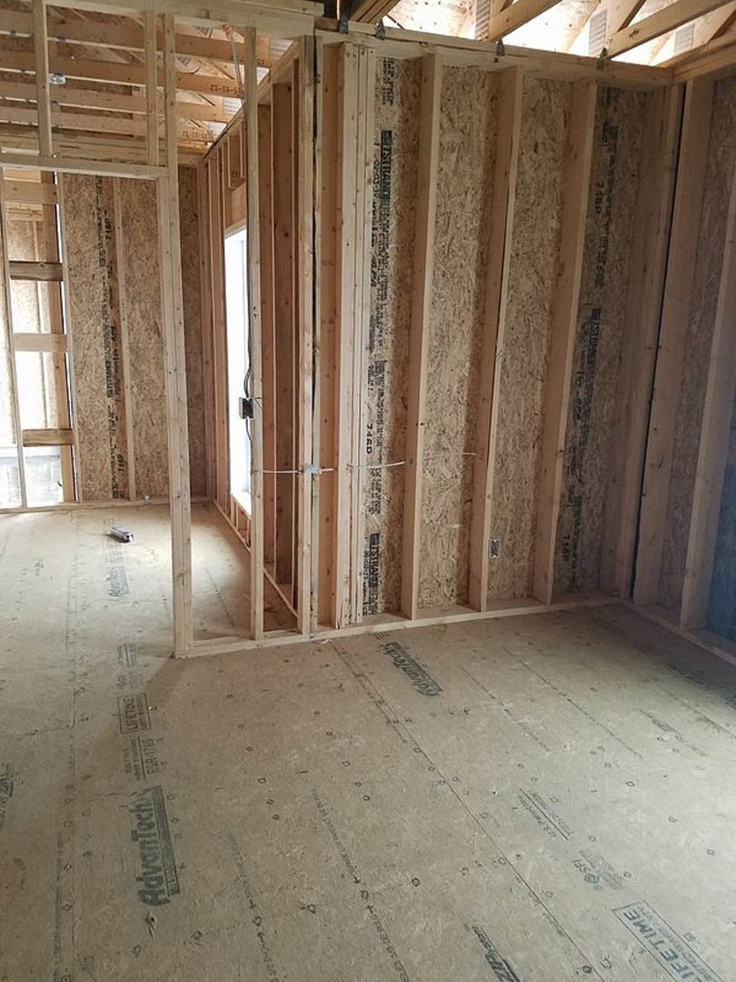 Room under construction with exposed wood framing, unfinished plaster walls, ceiling beams, and concrete floor; window installation visible.