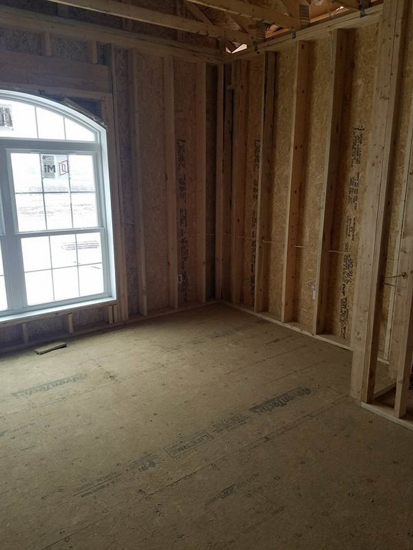 Room under construction with exposed wood framing, plaster walls, daylight streaming through window, and unfinished plank floor.