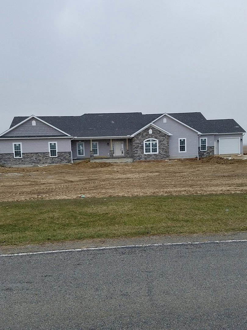 Partially built house with grey roof, white-framed windows, surrounded by grass and dirt road under cloudy sky