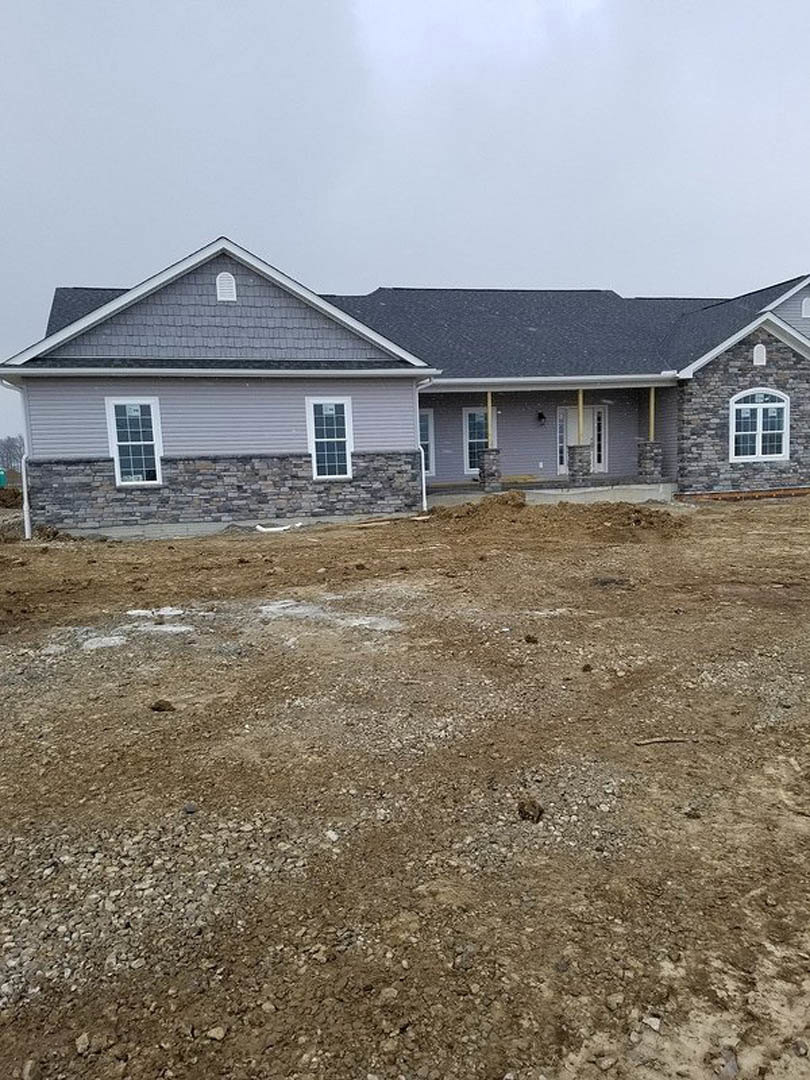 Two-story house under construction with grey roof, white-framed windows, and light siding, set behind a dirt field and brick retaining wall