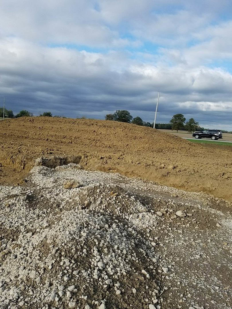 Dirt hill with scattered rocks in an open field, cloudy sky overhead, parked car and lone tree in the background
