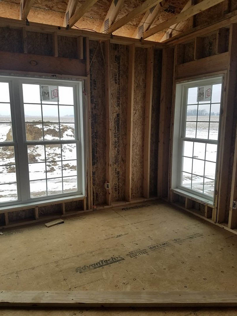 Room featuring wood plank walls, exposed beams, large windows with views of snowy ground and open field, black text on light-colored floor, cardboard and logo details visible