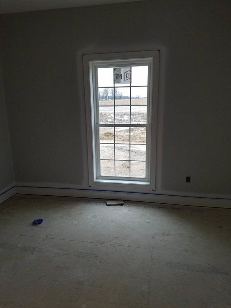 Large window set in a plaster wall, overlooking a grassy field, concrete flooring with a sheet of paper near the window, white ceiling, minimal interior fixtures.