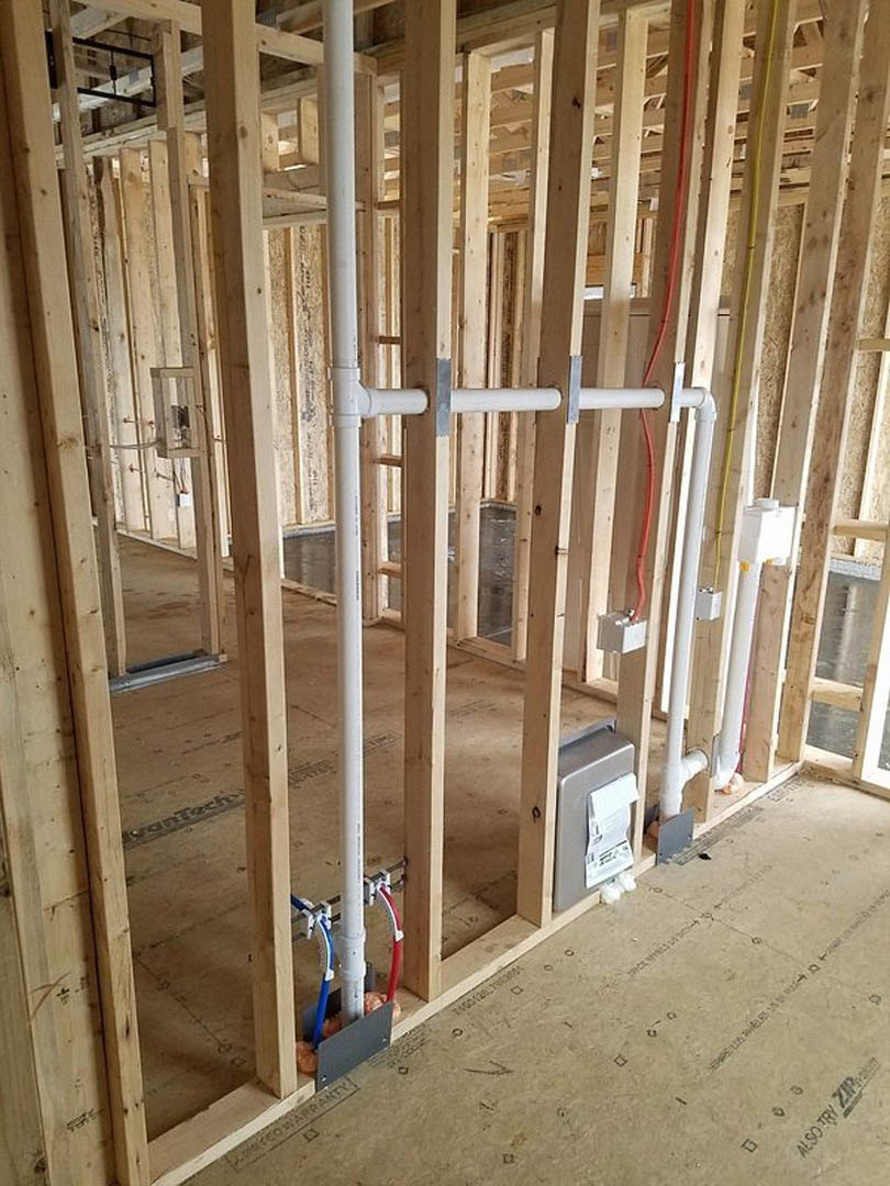 Unfinished room with exposed wooden framing, white pipes, red and blue hoses, electrical wiring, insulation, and a paper towel dispenser mounted on a wall.