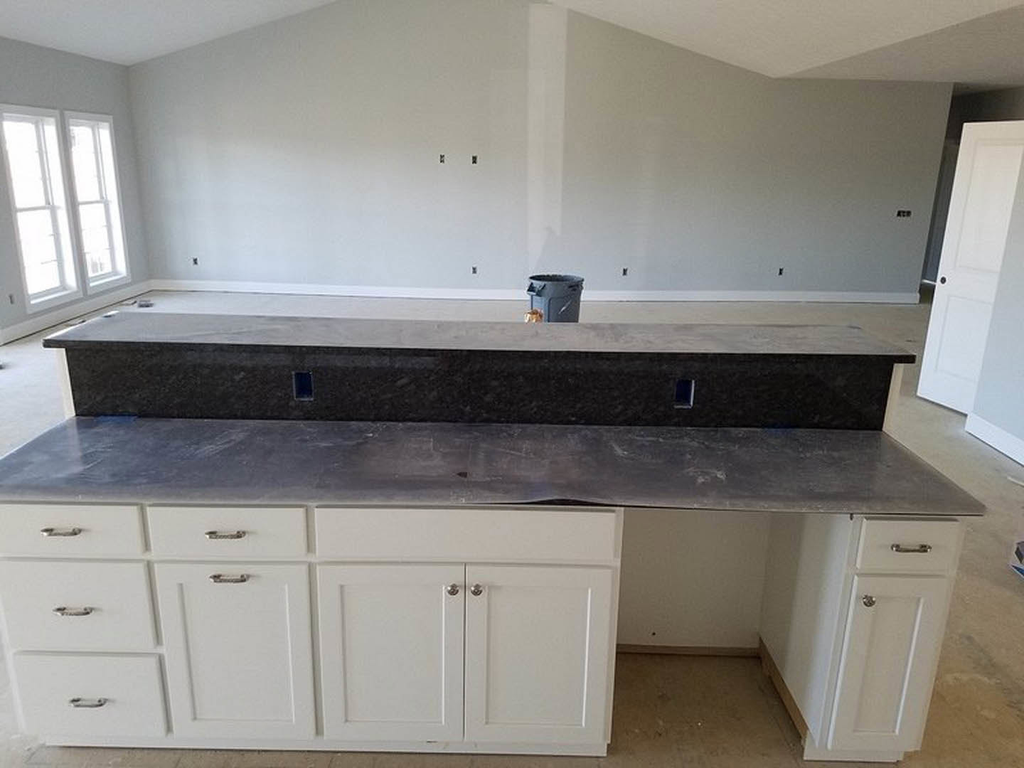 Granite kitchen countertop with tile backsplash, stainless steel sink, white cabinetry, wood beam detail, and black lidded trash can