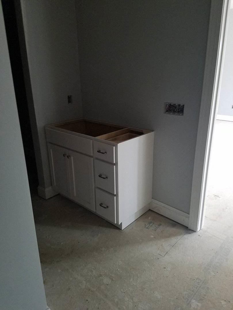 White shaker-style kitchen cabinet with silver handles against a plaster wall, wood flooring, and multiple drawers and cupboards.