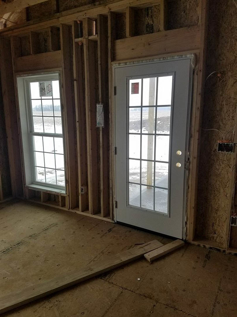 White-framed door and windows in a room with exposed wood framing, wooden plank flooring, and snowy landscape visible outside
