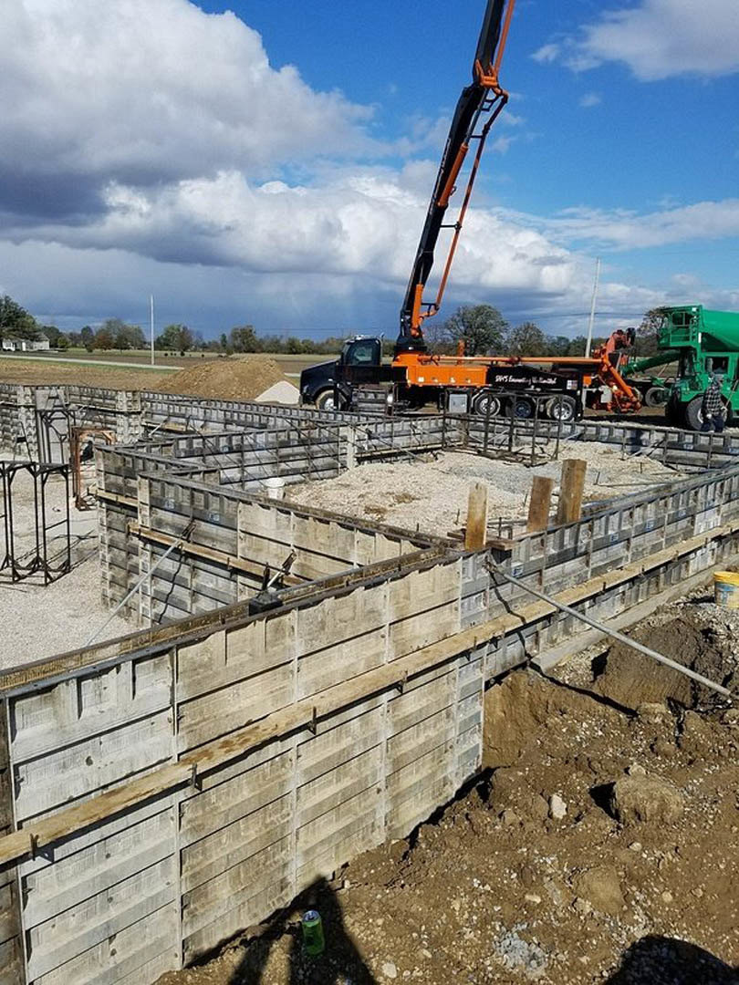 Framed foundation and piles of dirt on a residential construction site with a crane and green truck under a cloudy sky