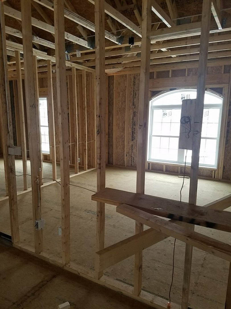 Exposed wood framing and beams in an unfinished room, plywood and lumber stacked on a table, white rectangular electrical box mounted on wall, rope tied to wooden board, black