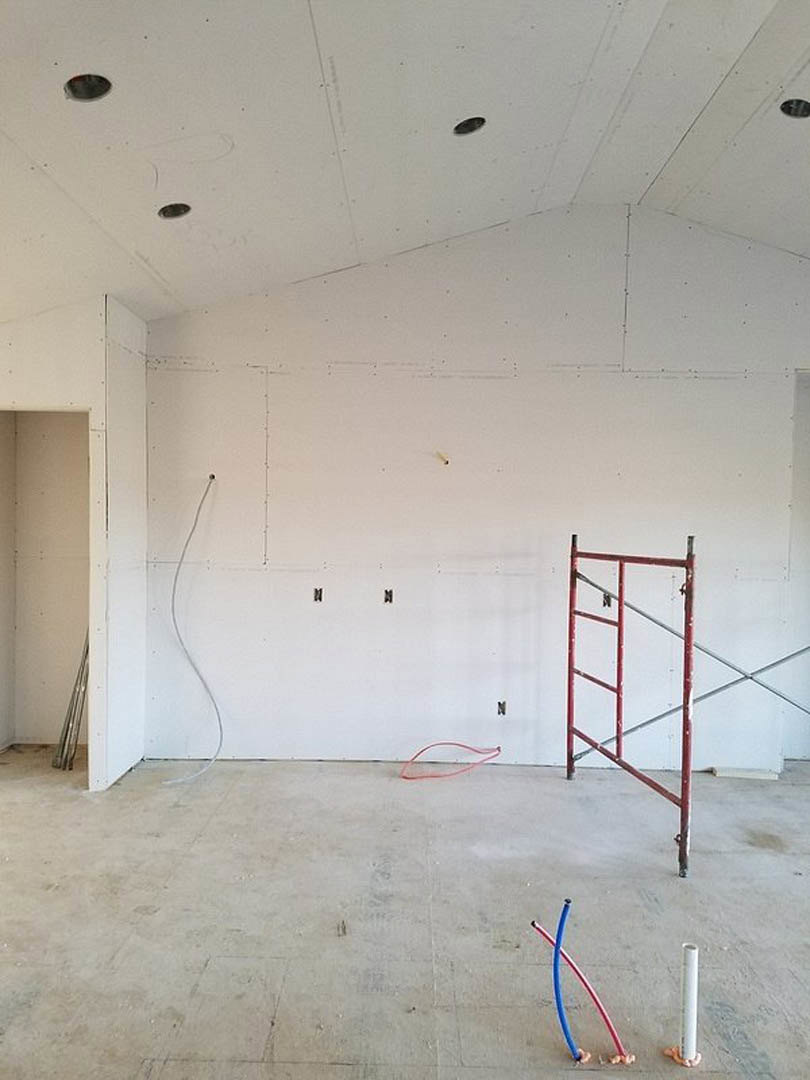 Unfinished room with white plaster walls, black trim, red scaffolding, blue and red hoses, ladder, and exposed ceiling under construction