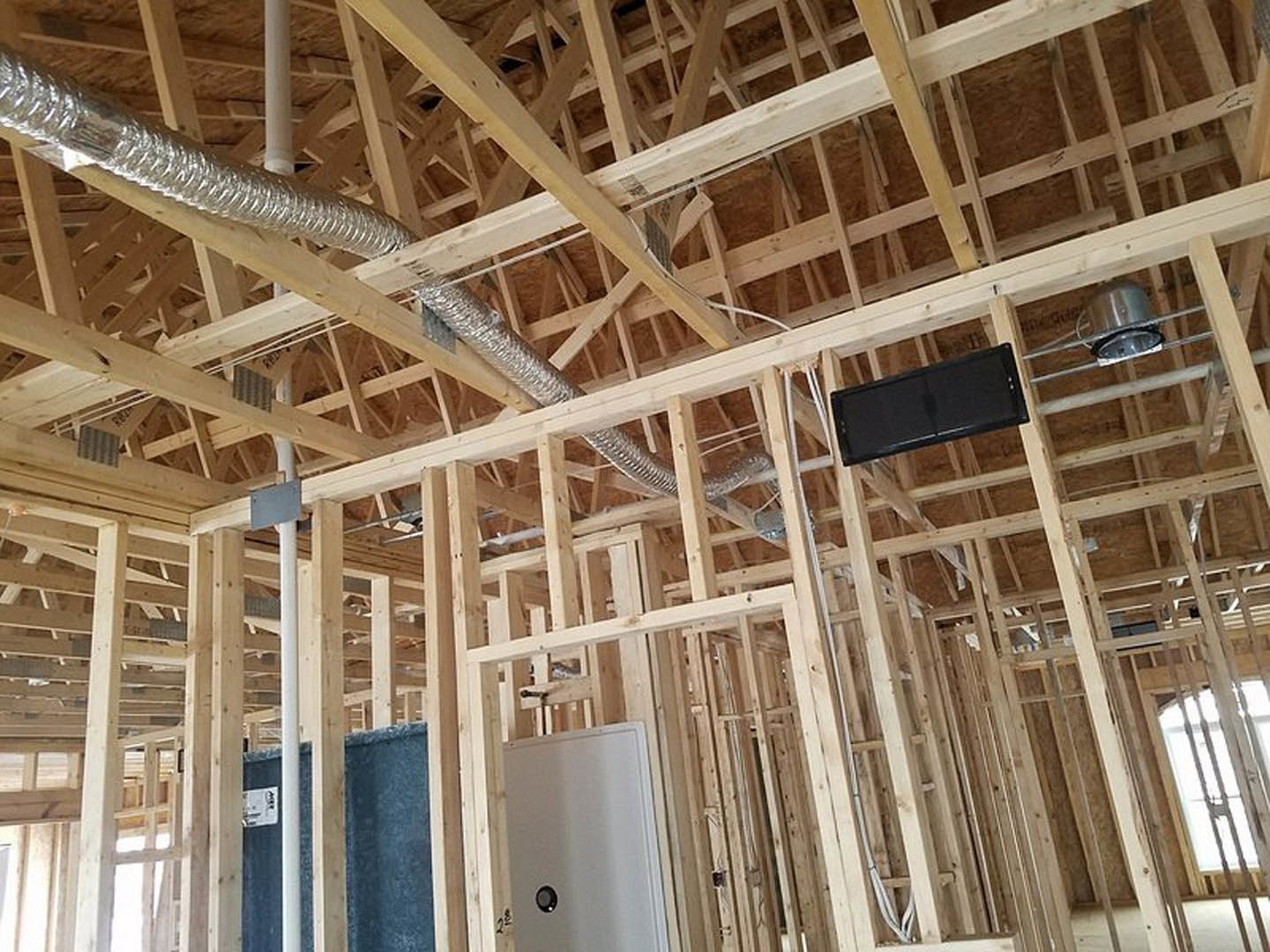 Exposed wood framing and beams inside a house under construction, with visible insulation, unfinished window opening, and construction materials scattered on plywood subfloor