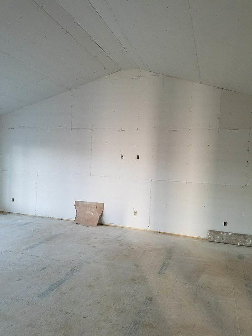 White plaster walls and ceiling with visible cracks, concrete floor partially covered by cardboard, broken tile near the center, exposed metal detail, unfinished and abandoned