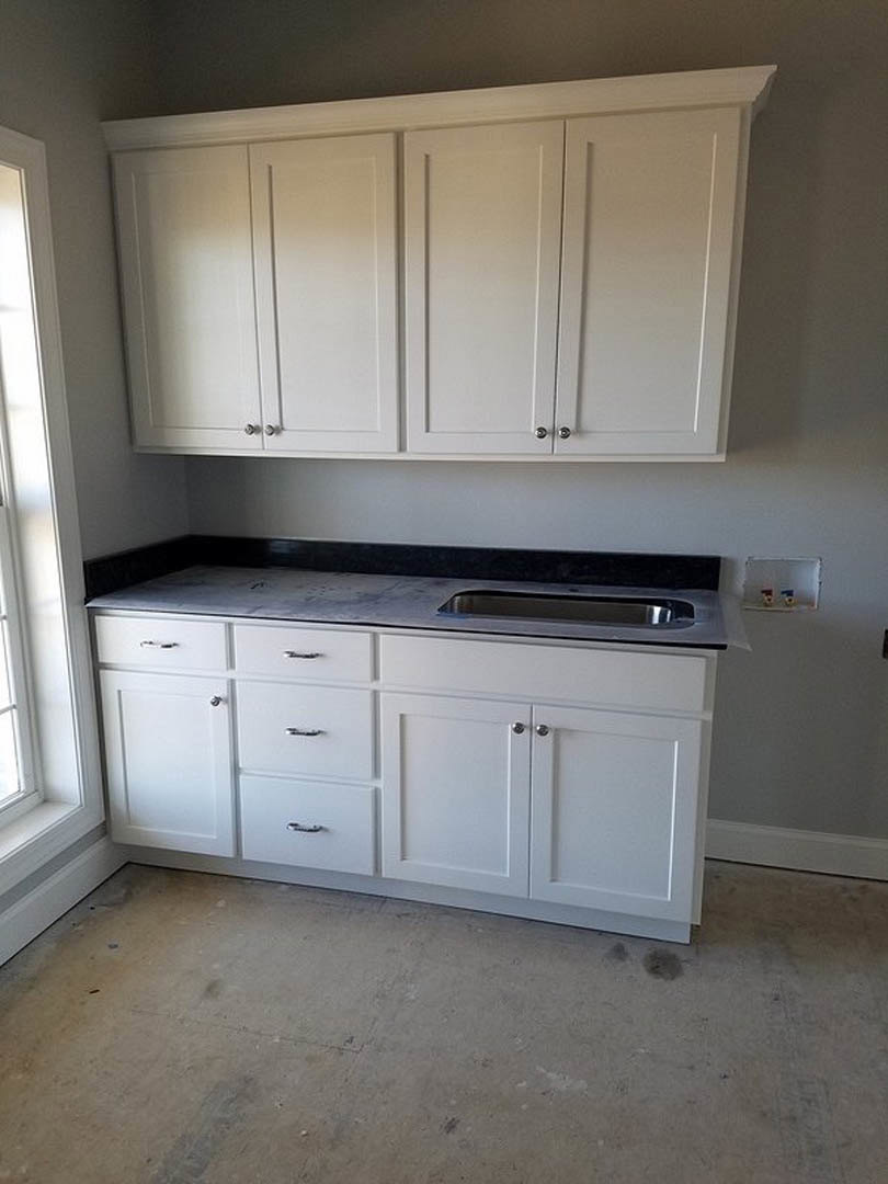 White kitchen with shaker cabinets and silver knobs, quartz countertop with undermount sink, stainless steel appliances, light wood flooring, sunlight streaming onto white walls.