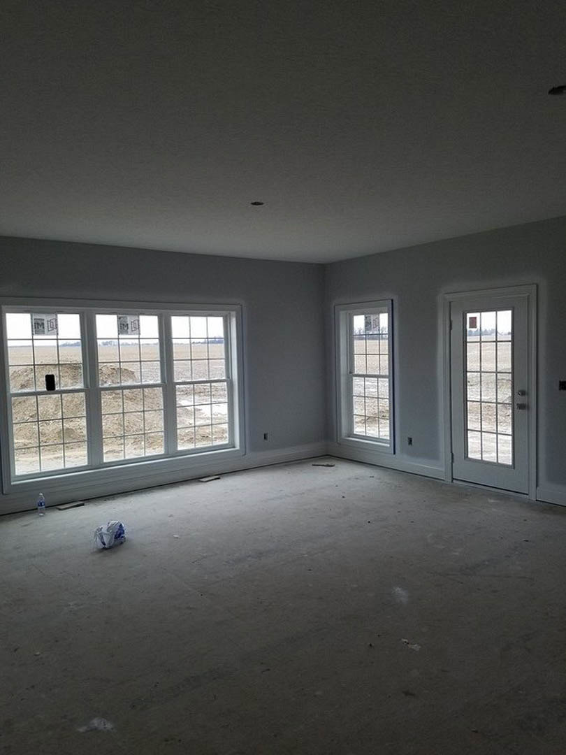 Bright room with large window overlooking a field, glass-paneled door, plaster walls, white furniture with blue accent, and unfinished floor.