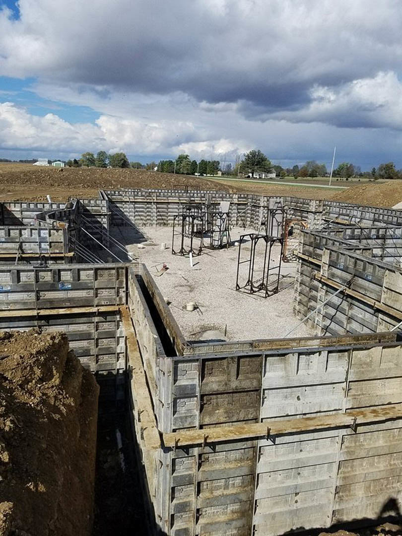 Expansive construction site with dirt mound, partially built concrete wall, metal framing on ground, and cloudy sky with scattered trees in background