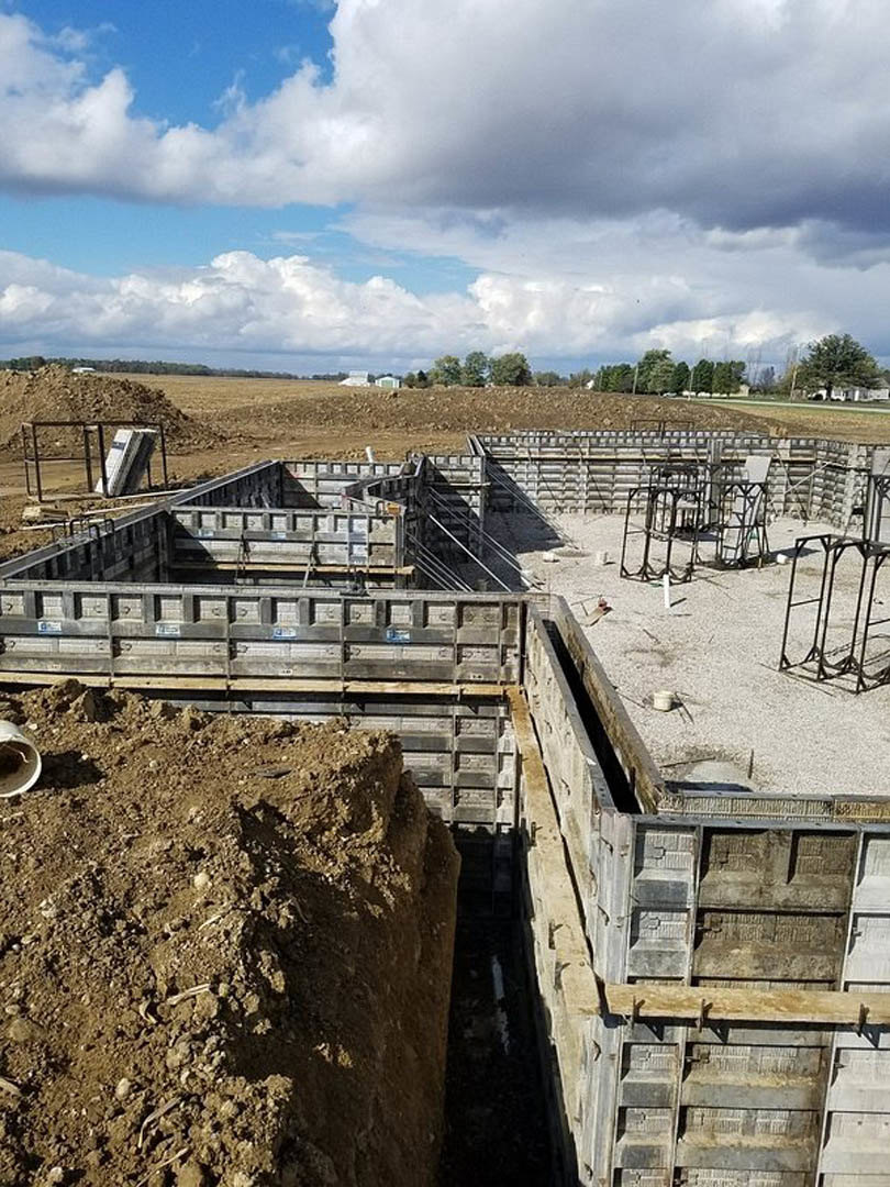 Construction site with exposed concrete foundation, piles of dirt, metal pipe, and unfinished wall under blue sky; trees visible in background