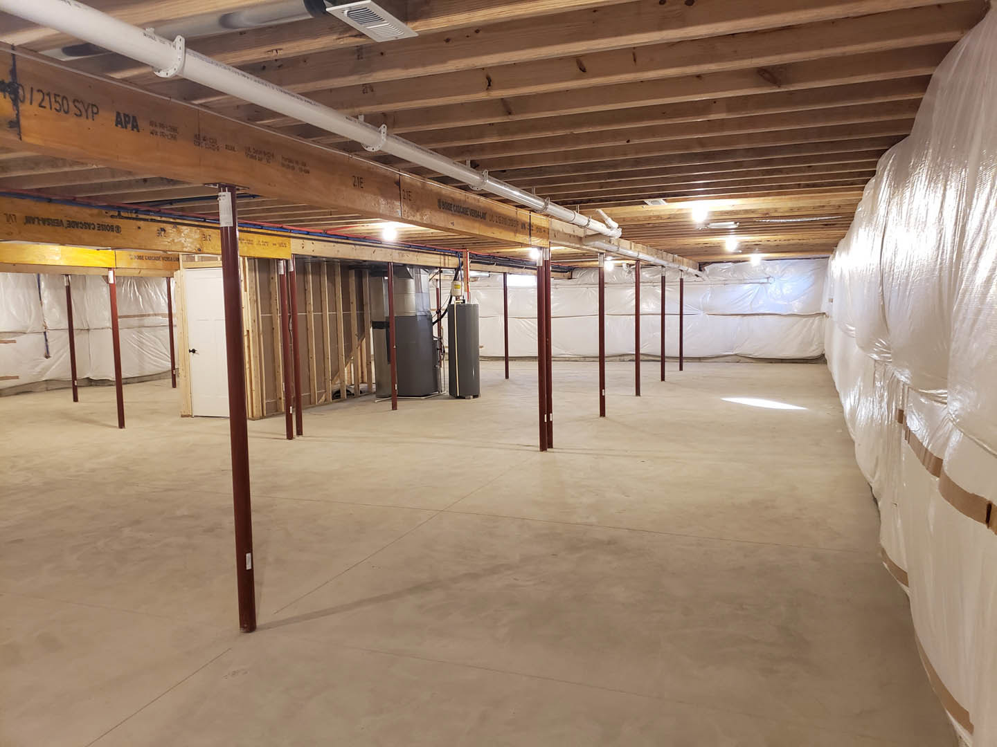 White-walled room with exposed wooden ceiling beams, visible pipes and vents, white plastic bags lined up on the floor, and a metal box mounted near the ceiling.