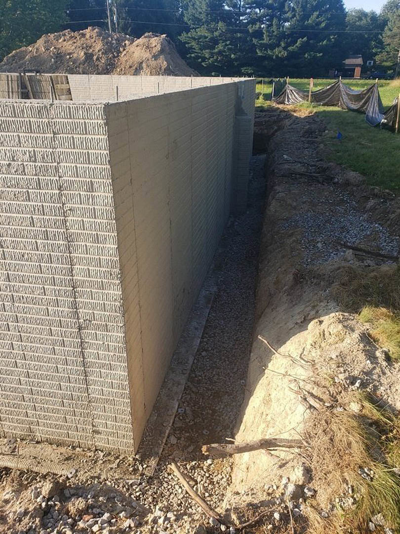Concrete exterior wall beside a dirt excavation, black tarp covering grass, tree trunk and wood debris in foreground, stone and composite materials visible