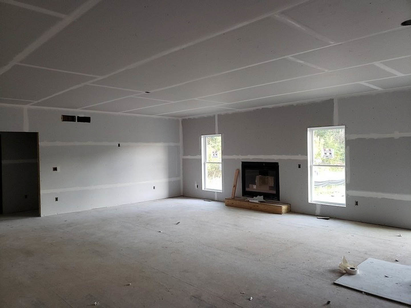 Living room with black fireplace featuring glass screen, large window with sign, plaster walls, and hardwood flooring; roll of toilet paper on floor.