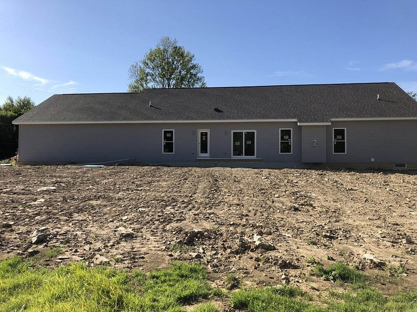 Grey house with white doors and glass windows, surrounded by a dirt field with patches of grass and a leafy green tree under a cloudy sky
