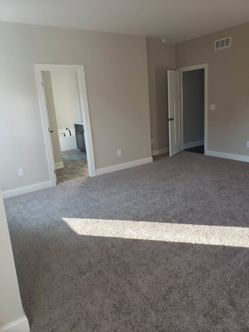 Carpeted bedroom with white walls and black trim, open door leading to bathroom featuring bathtub, sink, and wall vent
