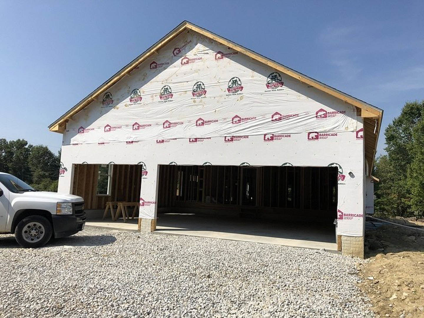 Partially built house with exposed framing and plastic sheeting, white pickup truck parked on dirt driveway, green leafy tree nearby, construction logo visible on truck door