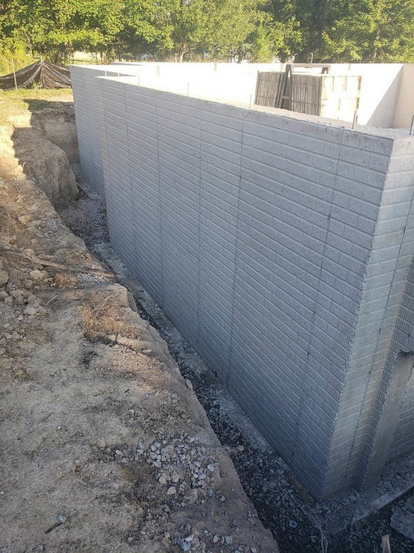 Concrete wall with circular hole in the ground, surrounded by grass, trees with green leaves, and a wooden gate set in a white wall; dirt and rocks adjacent to a brick wall.