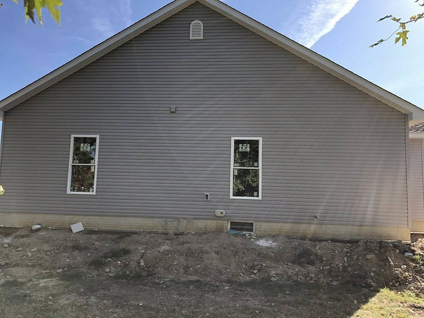 Grey siding house with white vent, large window displaying a plant and a sign, dirt patch on sloped yard under cloudy sky.