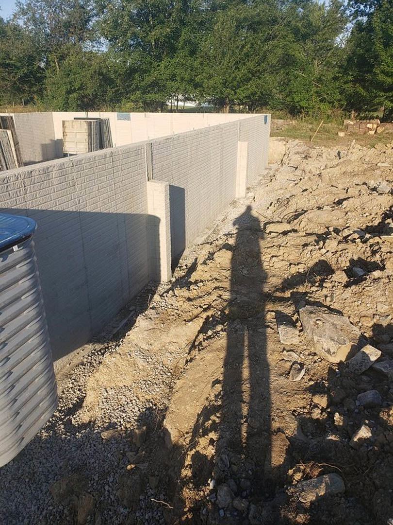 Shadow of a person cast on a dirt path near a group of trees, with a white door featuring several windows visible on a concrete foundation wall.