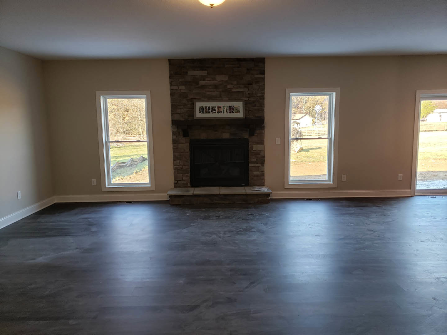 Living room with grey laminate flooring, white plaster walls, black-framed fireplace, large windows showing trees, fence, and neighboring house